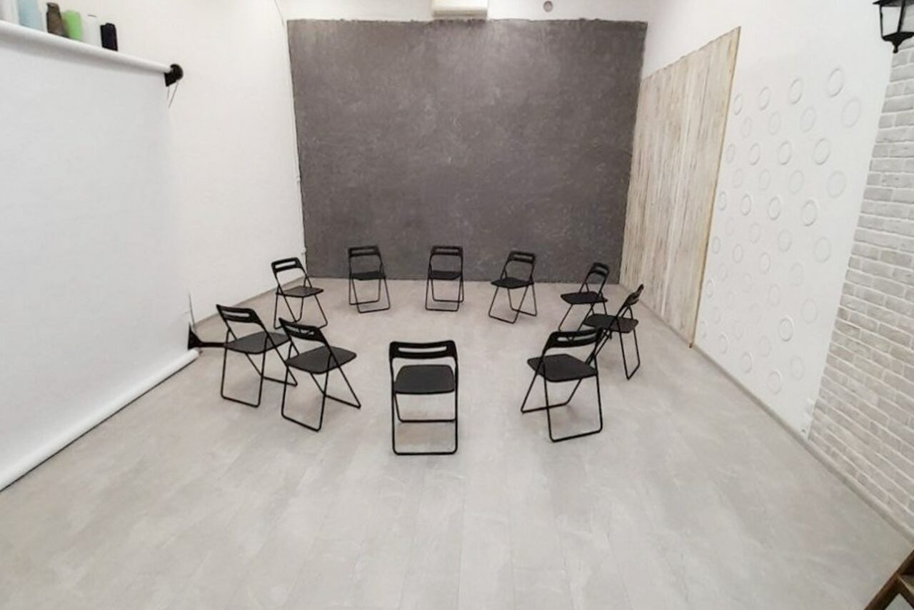 Арендовать Интер-Фото. Пространство с серыми стенами и светлым полом. Расставлены черные складные стулья по кругу. Справа белая стена с круглыми узорами, слева белый экран. Одна стена имитирует кирпич, другая - дерево. Освещение невидимо.