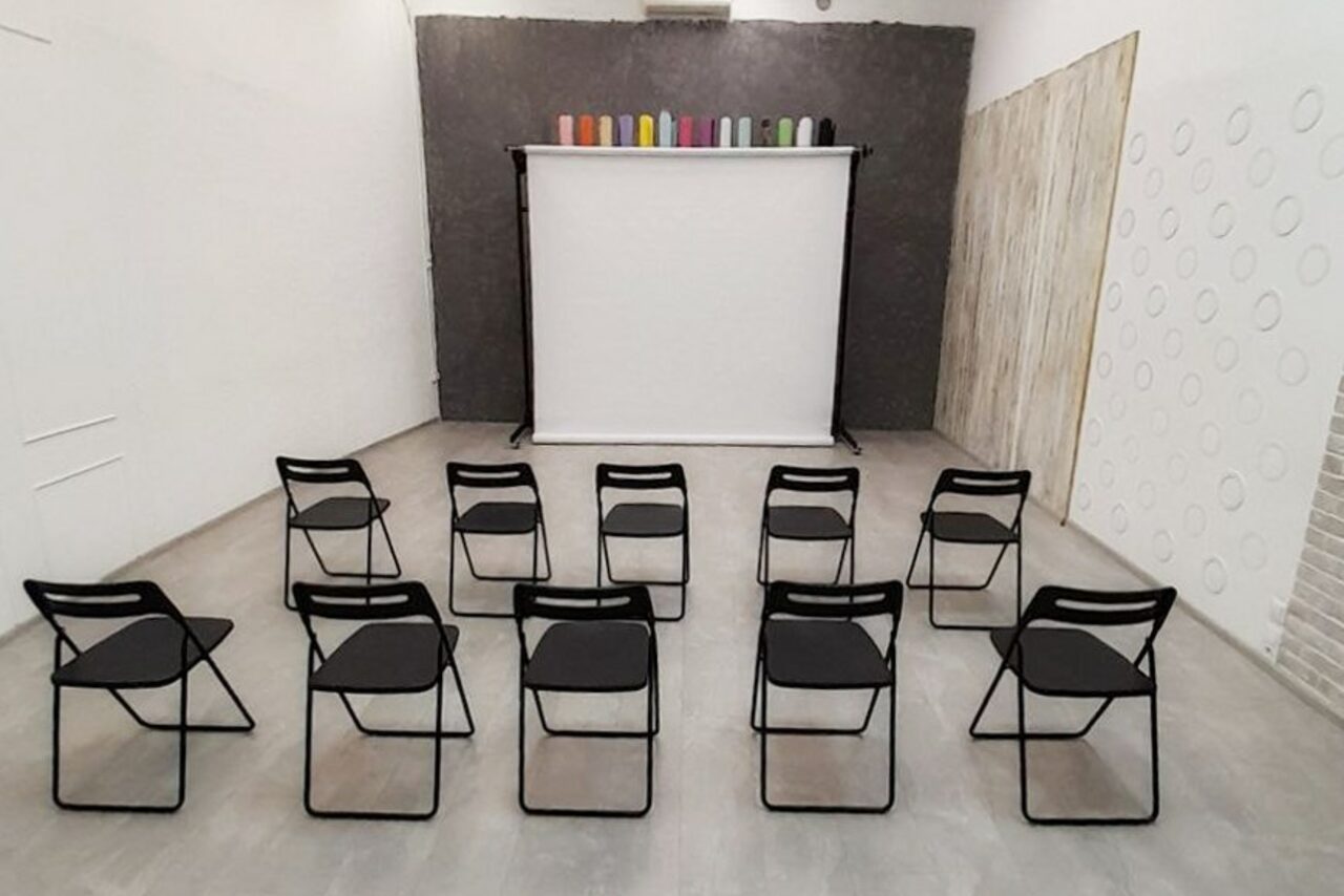 Арендовать Интер-Фото. Пространство с серыми стенами, пол из светлого материала. В центре 12 черных складных стульев, ориентированных к черно-белому экрану на штативе. Сверху экрана разноцветные цилиндры. Стена декорирована линейными и круглыми элементами.