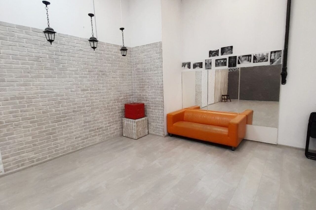 Арендовать Интер-Фото. Студийное помещение с кирпичными стенами светло-серого оттенка. Оранжевый диван, большое зеркало, чёрно-белые фотографии на стене. Лампы подвесные чёрные. Полы светло-серые. Корзина с красным покрышкой. Несколько стульев.