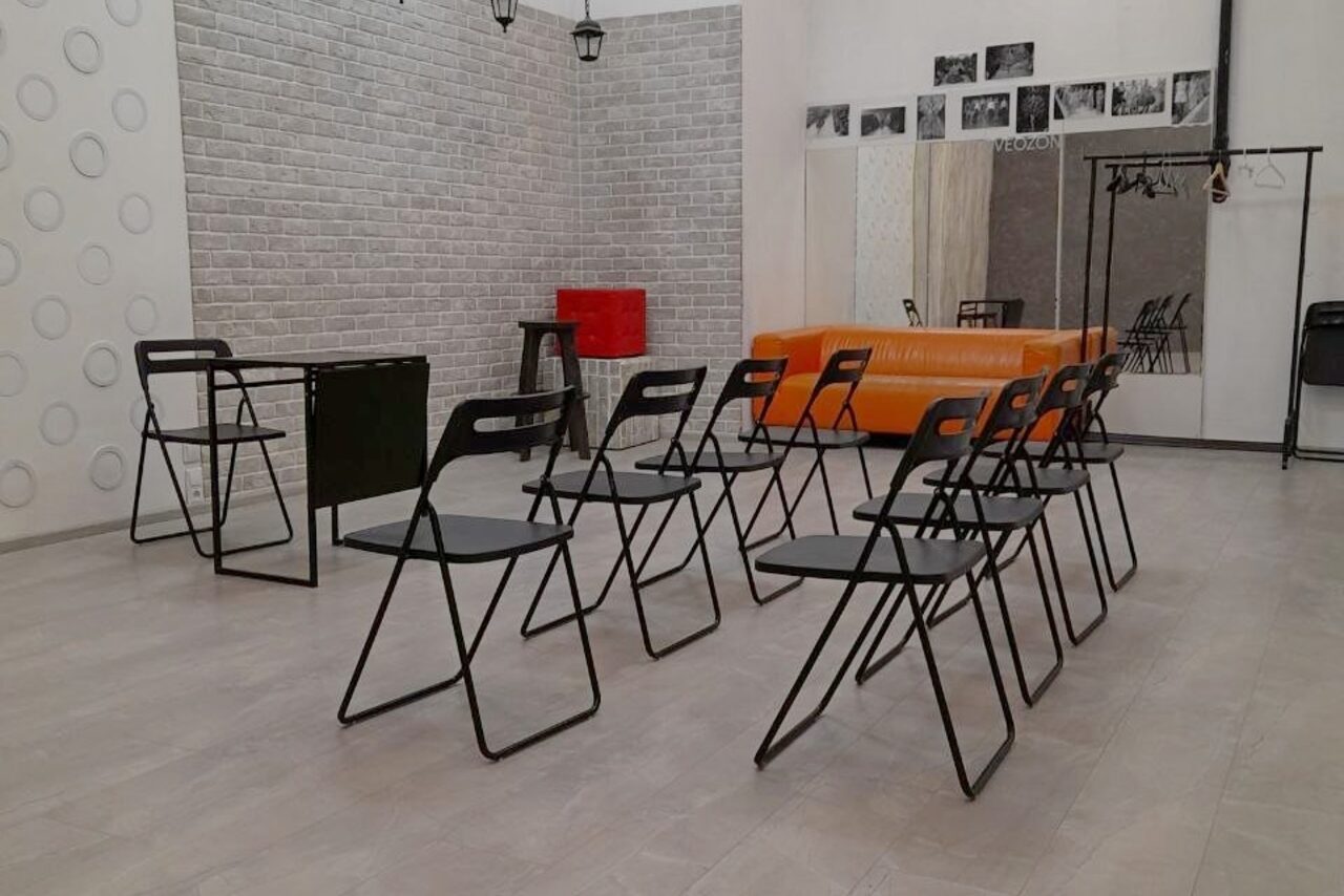 Арендовать Интер-Фото. Помещение с рядной расстановкой черных складных стульев, черный стол напротив. Оранжевый диван у стены. Белая кирпичная стена с узором из кругов и черно-белыми фотографиями. Металлическая вешалка. Светло-серый пол.