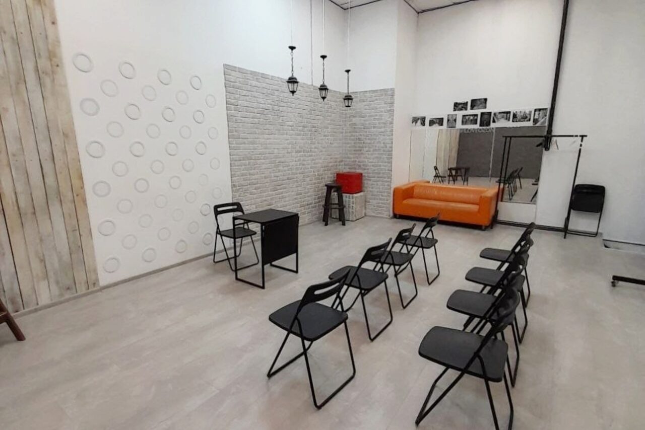 Арендовать Интер-Фото. Помещение с деревянным полом, стульями в ряд, оранжевым диваном, столом и табуретом, декоративной стеной с кругами. На стенах фото и зеркало. Подвесные светильники, светлый и кирпичный декор стен, стойка для одежды.