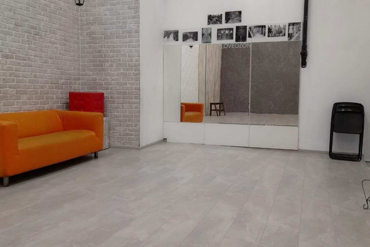 Арендовать Интер-Фото. Просторное помещение с белыми и серыми стенами, частично отделанными кирпичной кладкой. На полу светлый ламинат. Оранжевый диван, красный куб, черный стул. Зеркальная стена с фотографиями сверху. Черная труба вдоль стены. Надпись "LOVEZONE".