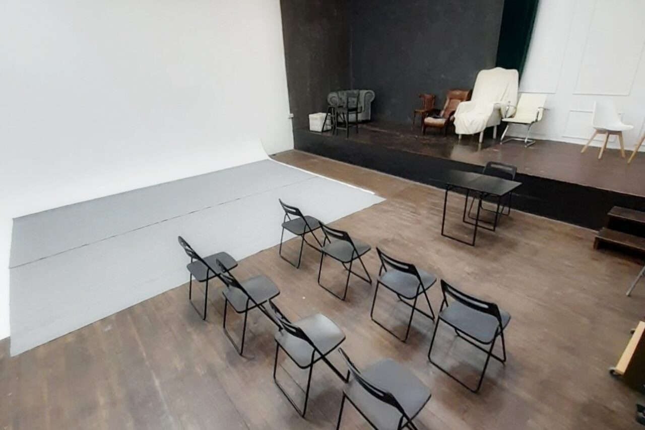 Арендовать Интер-Фото. Студийное пространство с белой и черной стенами, деревянные полы. Несколько стульев из черного пластика, стол. Мягкие кресла в углу на подиуме, частично накрыты тканью. Освещение отсутствует.
