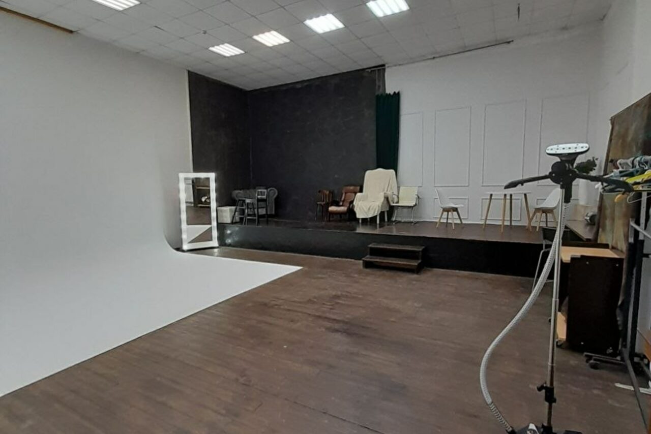 Арендовать Интер-Фото. Просторное помещение с деревянным полом, белой и черной фотозонами. Подиум с креслами и стульями покрытыми тканью. Освещение от потолочных ламп. На переднем плане фототехника на стойке. Стены белые с декоративной отделкой, одна черная.