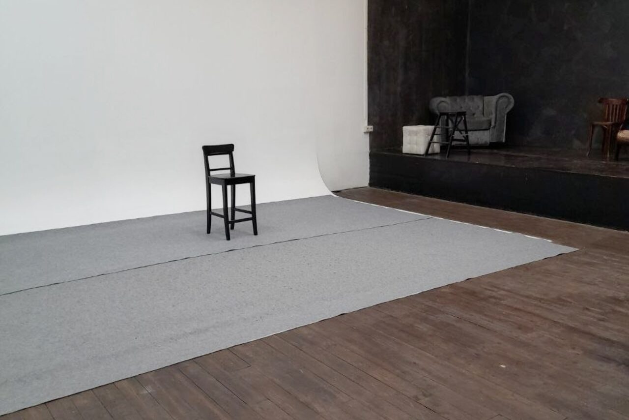 Арендовать Интер-Фото. Студийное пространство с гладким белым фоном. В центре черный стул на сером ковре. Справа подиум из темного дерева с серым диваном и подушками, позади черная и белая стена. Освещение отсутствует. Пол из деревянных досок.