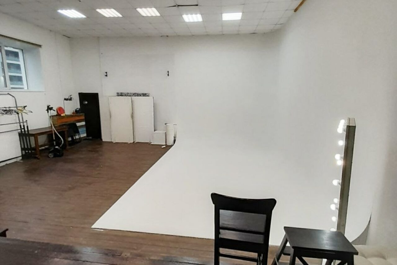 Арендовать Интер-Фото. Студийное пространство с циклорамой, темный пол, белые стены и потолок, сетчатые светильники, рабочий стол, стул, зеркало с лампами, окно с решеткой справа, несколько роликов бумаги у стены.