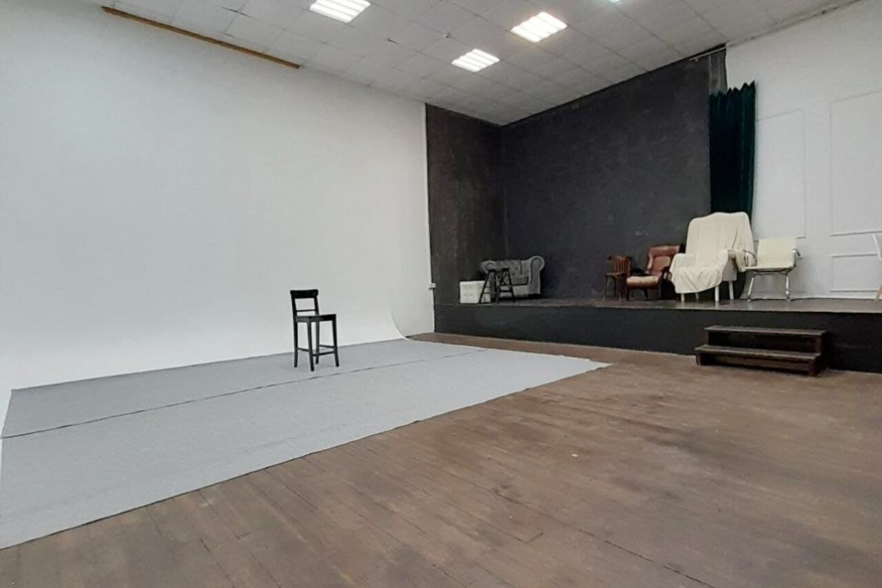 Арендовать Интер-Фото. Просторное помещение с циклорамой, деревянный пол, серые стены. Один черный стул на циклораме. Подиум с креслами, диваном и покрытым чехлом креслом. Потолочные светильники. Серые и белые цвета, лаконичный интерьер.