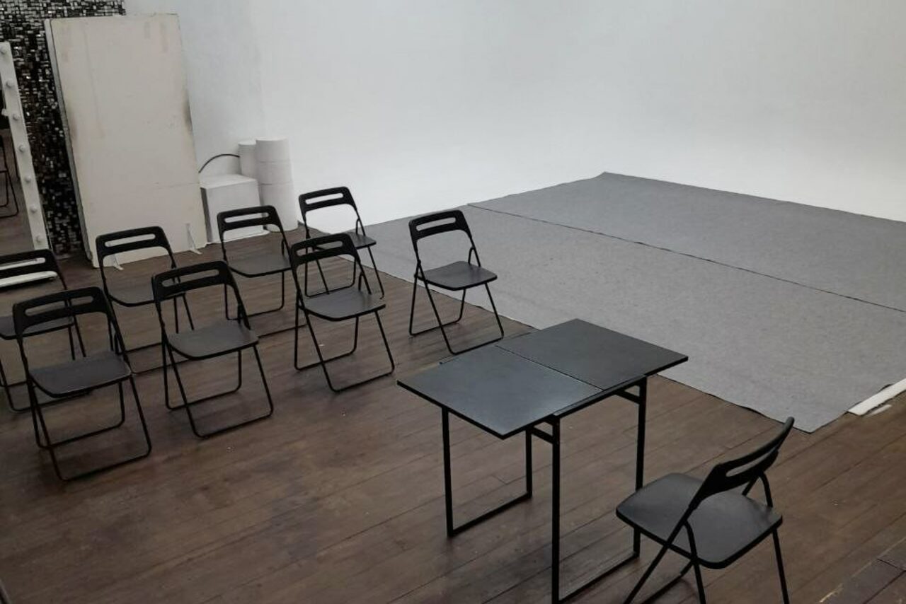 Арендовать Интер-Фото. Пространство со светлыми стенами и ковровым покрытием. Расставлены черные металлические стулья и два стола. На полу темное деревянное покрытие. В углу белая дверь и свернутые рулоны бумаги.