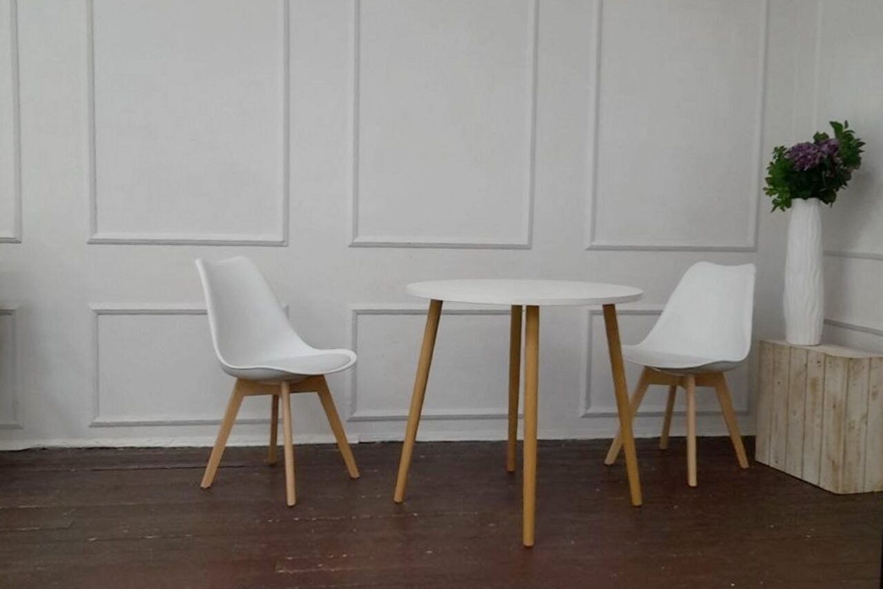 Арендовать Интер-Фото. Пространство с деревянным полом, светлыми стенами с панелями. В центре круглый стол с белой поверхностью и деревянными ножками, окружён двумя белыми стульями с деревянными ножками. Справа белая ваза с зеленью и фиолетовыми цветами на деревянной тумбе.