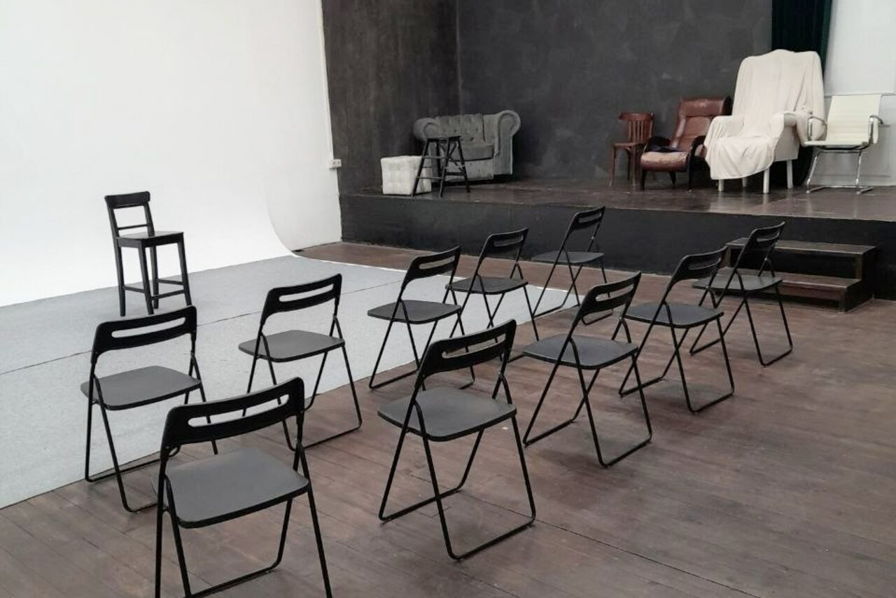 Арендовать Интер-Фото. Пространство с затемненной сценой и зрительным залом. На сцене диван и кресла. В зале ряды черных стульев, деревянный пол. Стены темные, пол светлый. Освещение отсутствует.