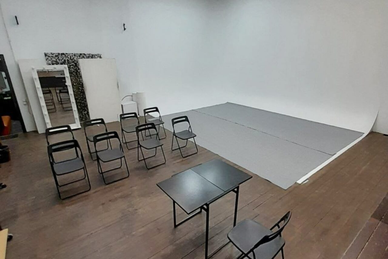 Арендовать Интер-Фото. Фотостудия с белым циклическим фоном, черными складными стульями в линию, двумя черными столами, бежевым деревянным полом. Светлые стены, крупное зеркало у стены, серый ковролин на фоне. Светлое однотонное освещение.