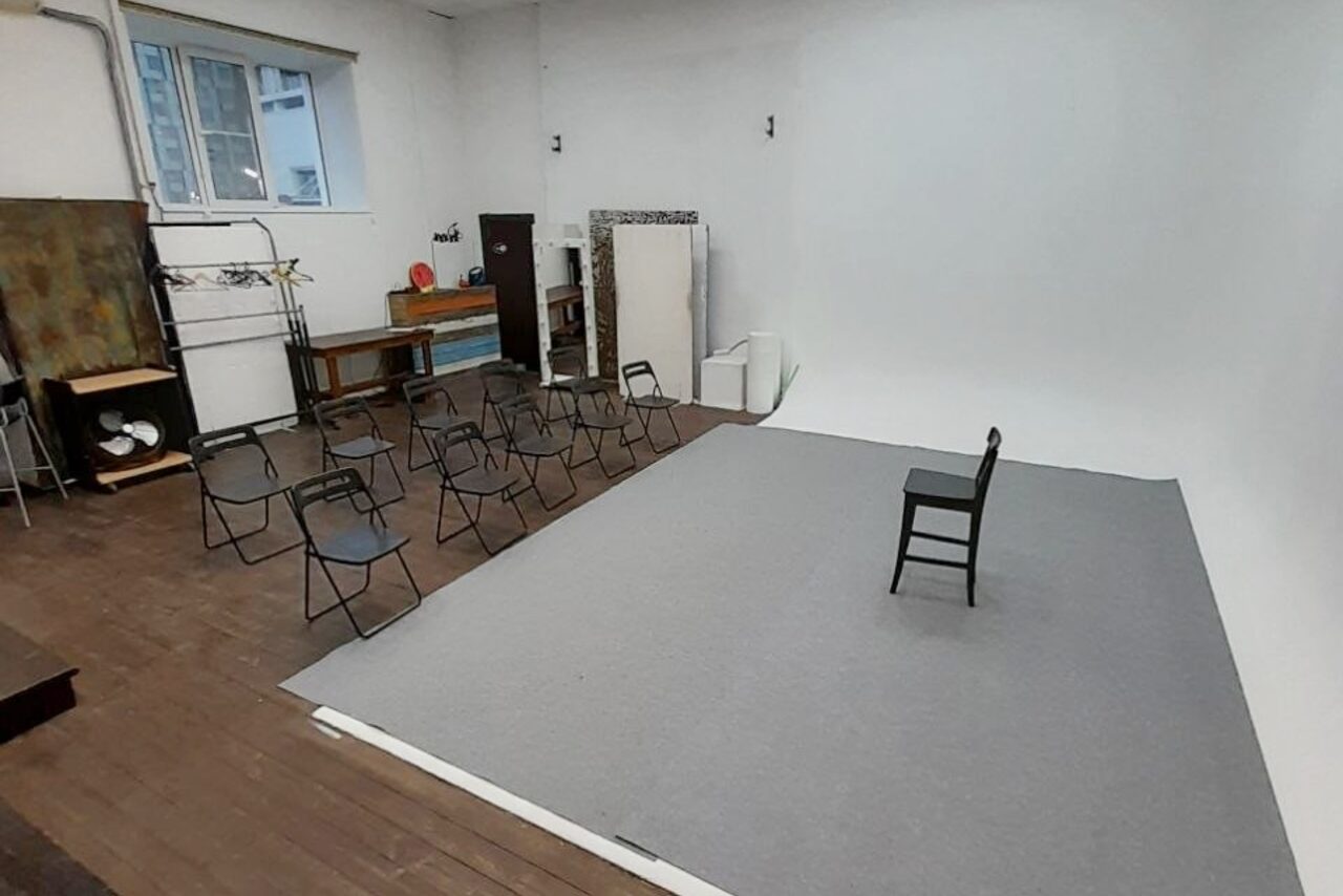 Арендовать Интер-Фото. Студийное помещение с циклорамой; справа серый коврик, черный стул; слева складные стулья, 10 штук; деревянный пол, белые стены; на заднем плане столы, лампы, оборудование; большое окно; белая дверь.