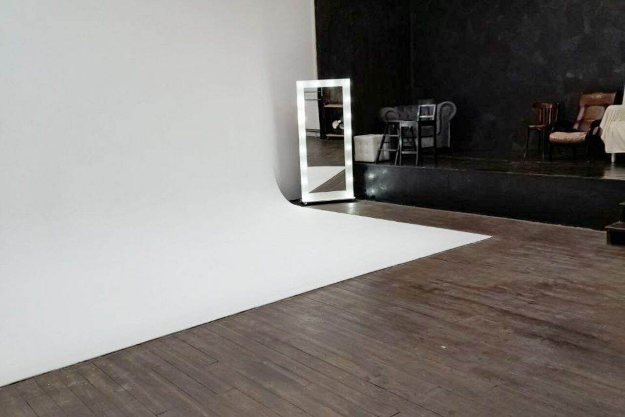 Арендовать Интер-Фото. Просторное помещение с фотостудией, включает белый циклораму и темные деревянные полы. На заднем плане чёрная стена, кресла, стул и стол. Установлено большое зеркало с подсветкой. Освещение неяркое, материалы — дерево и текстиль.