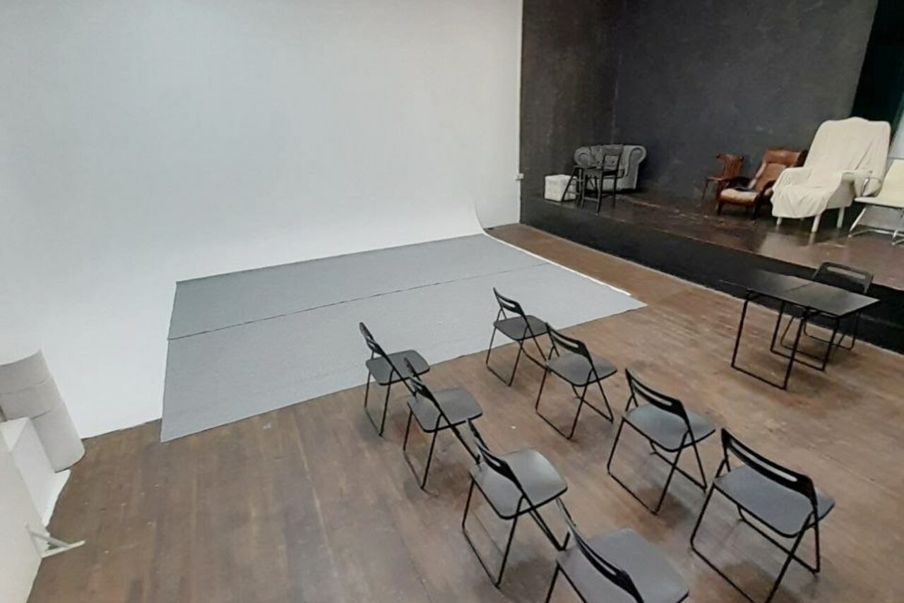 Арендовать Интер-Фото. Студийное пространство с деревянным полом, светлыми стенами и обтекаемым фоном для съемок. Несколько металлических стульев рядами, стол, в углу — зона с диваном и креслами. Освещение отсутствует, мебель простая, приглушенные цвета.