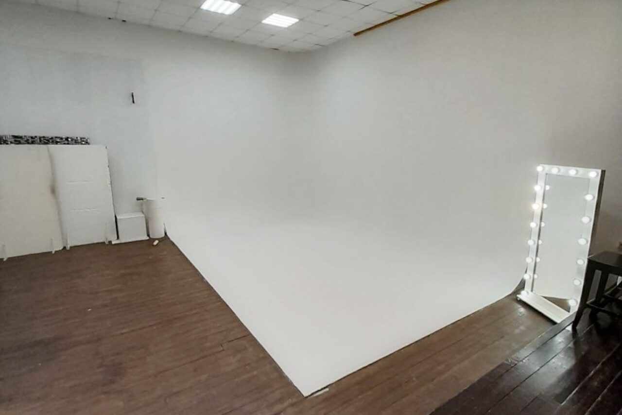 Арендовать Интер-Фото. Студийное пространство с белым циклом и темным деревянным полом. В правой части стоит зеркало с лампочками по периметру. На заднем плане сложенные белые панели. Потолок с квадрaтными светильниками.