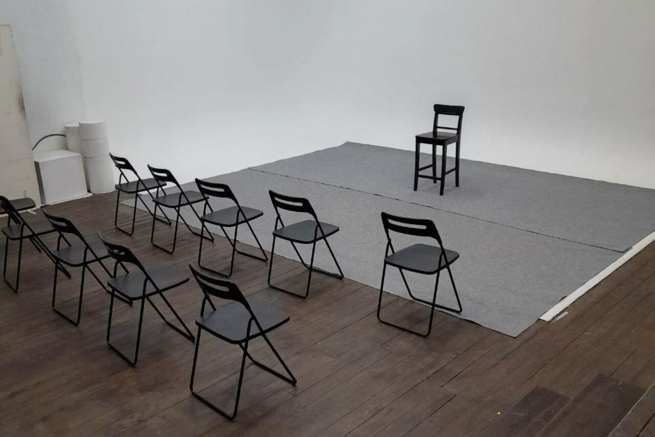 Арендовать Интер-Фото. Пространство с деревянным полом и белыми стенами, оснащено черными складными стульями, расположенными в два ряда, перед серым ковром на возвышении, где стоит высокий стул. Освещение нейтральное, справа видны белые цилиндрические объекты.