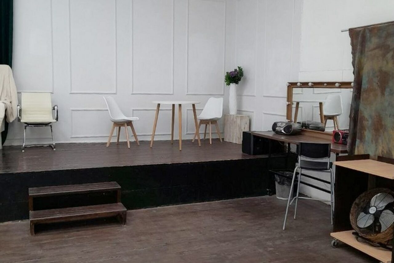 Арендовать Интер-Фото. Многофункциональное пространство с деревянной сценой, двумя белыми стульями и столиком. Зеркало с лампочками на тумбе, рядом стол с аудиотехникой. Отделка светлыми панелями, тёмный пол. Рядом ширма со светлым и тёмным участками, сбоку стоит вентилятор.