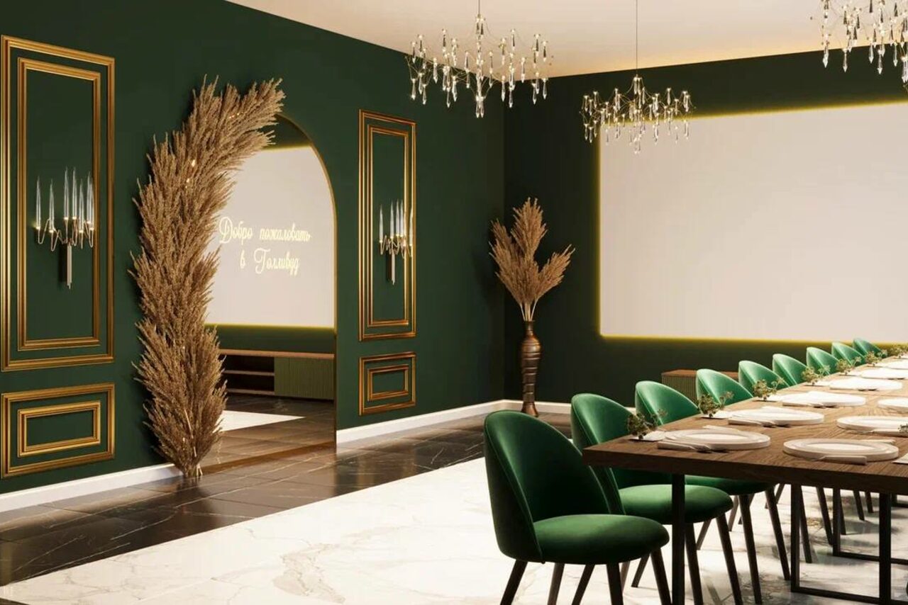 Арендовать Голливуд. Элегантный лофт в зелено-бежевых оттенках. Пространство с длинным столом и зелёными стульями. Тёмно-зелёные стены с золотыми рамками. На полу бело-коричневый мрамор. Подвесные люстры, неоновая надпись, декоративные растения. Арочный проход. Светлое потолочное освещение.