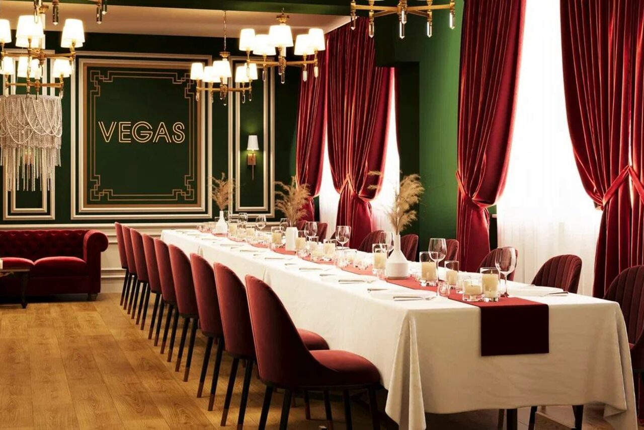 Арендовать Вегас. Атмосферный лофт с акцентным дизайном. Просторное помещение с длинным банкетным столом, покрытым белой скатертью. Красные стулья, зеленые стены, надпись "VEGAS", позолоченные светильники и красные шторы. Пол деревянный, свет из окон и люстр, украшения из сухих растений.