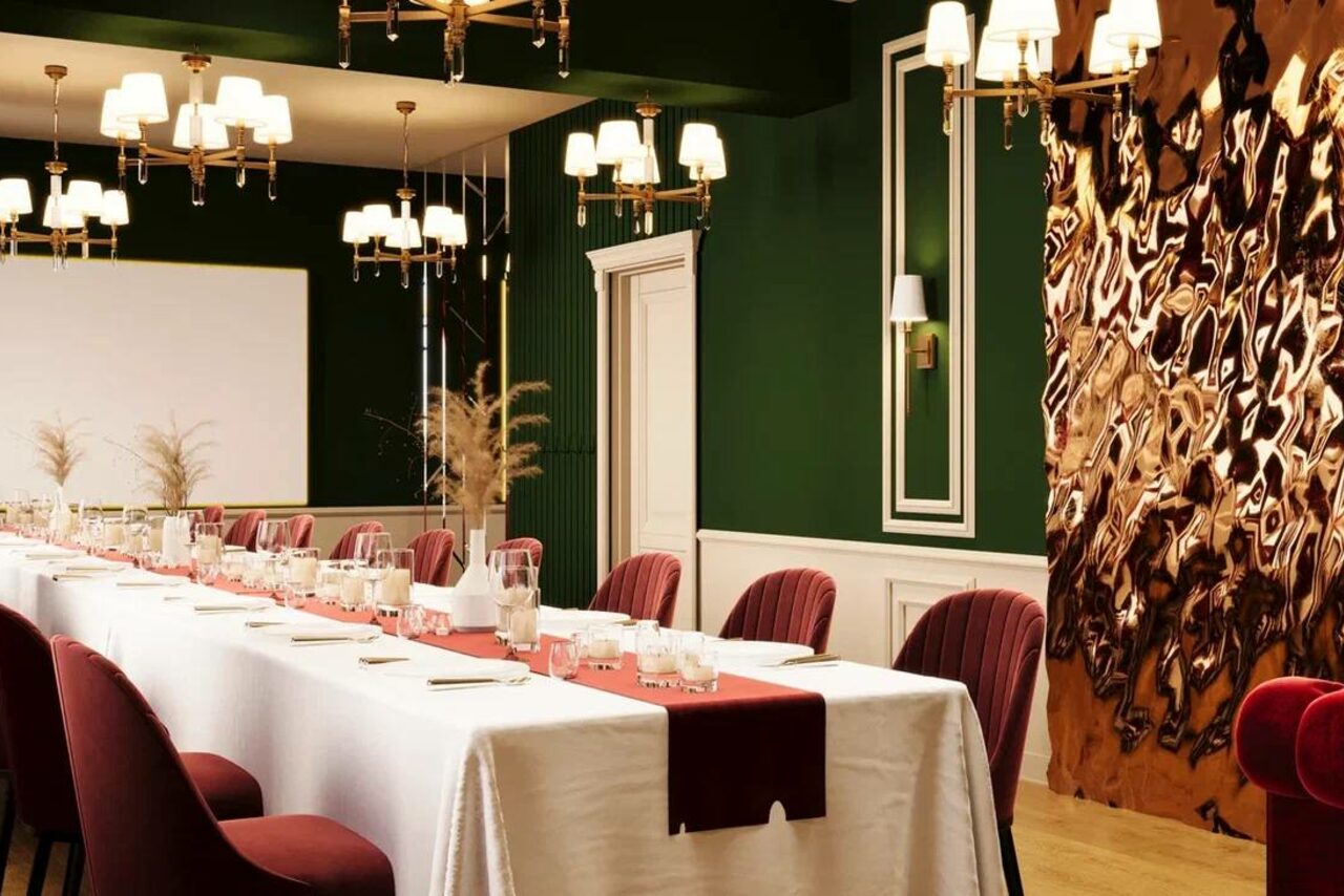 Арендовать Вегас. Атмосферный лофт с акцентным дизайном. Просторная комната с длинным столом, накрытым белой скатертью и бордовыми сервировочными дорожками. Вокруг бордовые стулья. Тёмно-зелёные стены, золотые и белые светильники, декоративная панель с текстурой. Светлое деревянное покрытие пола.