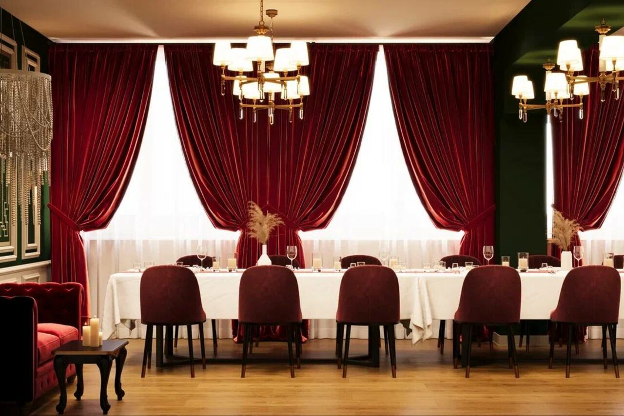 Арендовать Вегас. Атмосферный лофт с акцентным дизайном. Пространство для мероприятий с длинным столом, покрытым белой скатертью; красные бархатные занавески, стулья бордового цвета, люстры с лампами, деревянный пол, бокалы на столе, свечи на маленьком столике, зеленые стены.