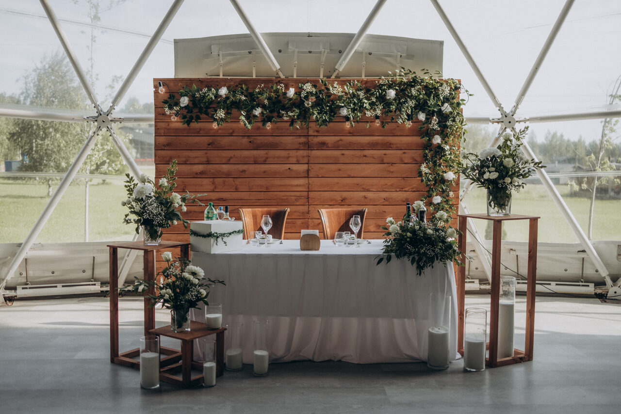 Арендовать Эко-отель "Джао Да!Ча". Шатер-Сфера. Шатровое помещение с треугольными окнами, деревянная стена с гирляндой из зелени и цветов, белый стол с белой скатертью, стеклянные декоративные элементы, свечи, бокалы, столовые приборы, декорации из живых растений.