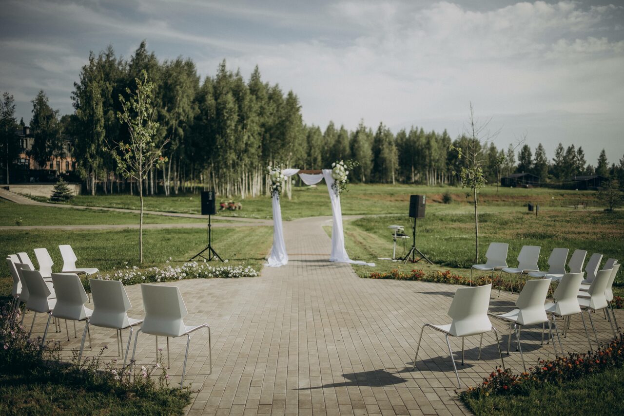 Арендовать Эко-отель "Джао Да!Ча". Шатер-Сфера. Открытое пространство для мероприятий на улице, с каменной дорожкой, расположенной между двумя рядами белых стульев. В центре арка с белой тканью и цветами. Вокруг травяной газон и деревья. Установлены две колонки на стойках.