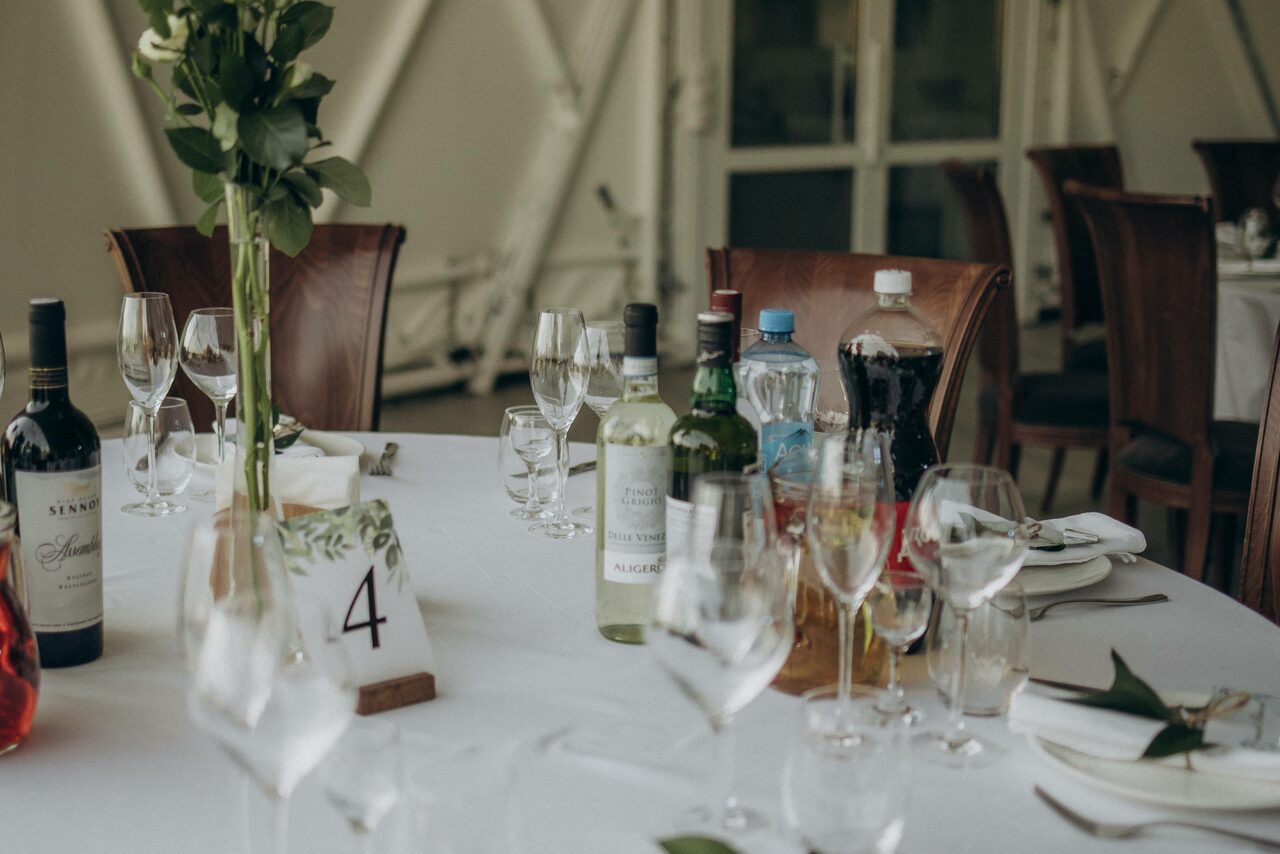 Арендовать Эко-отель "Джао Да!Ча". Шатер-Сфера. Круглый банкетный стол с белой скатертью, на котором стоит номер стола 4. На столе расставлены бутылки с вином и напитками, бокалы, тарелки с салфетками. Деревянные стулья вокруг стола. Фон из светлых стен и стеклянных дверей.