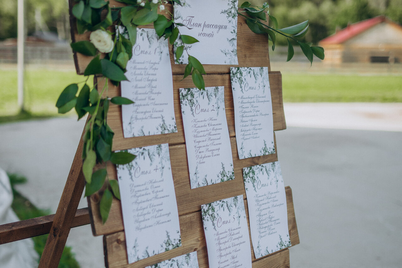 Арендовать Эко-отель "Джао Да!Ча". Шатер-Сфера. Деревянная стойка с прикрепленными карточками, украшенными зеленью. Карточки содержат текст и оформлены в виде плана рассадки гостей. На заднем плане видна открытая площадка и газон.