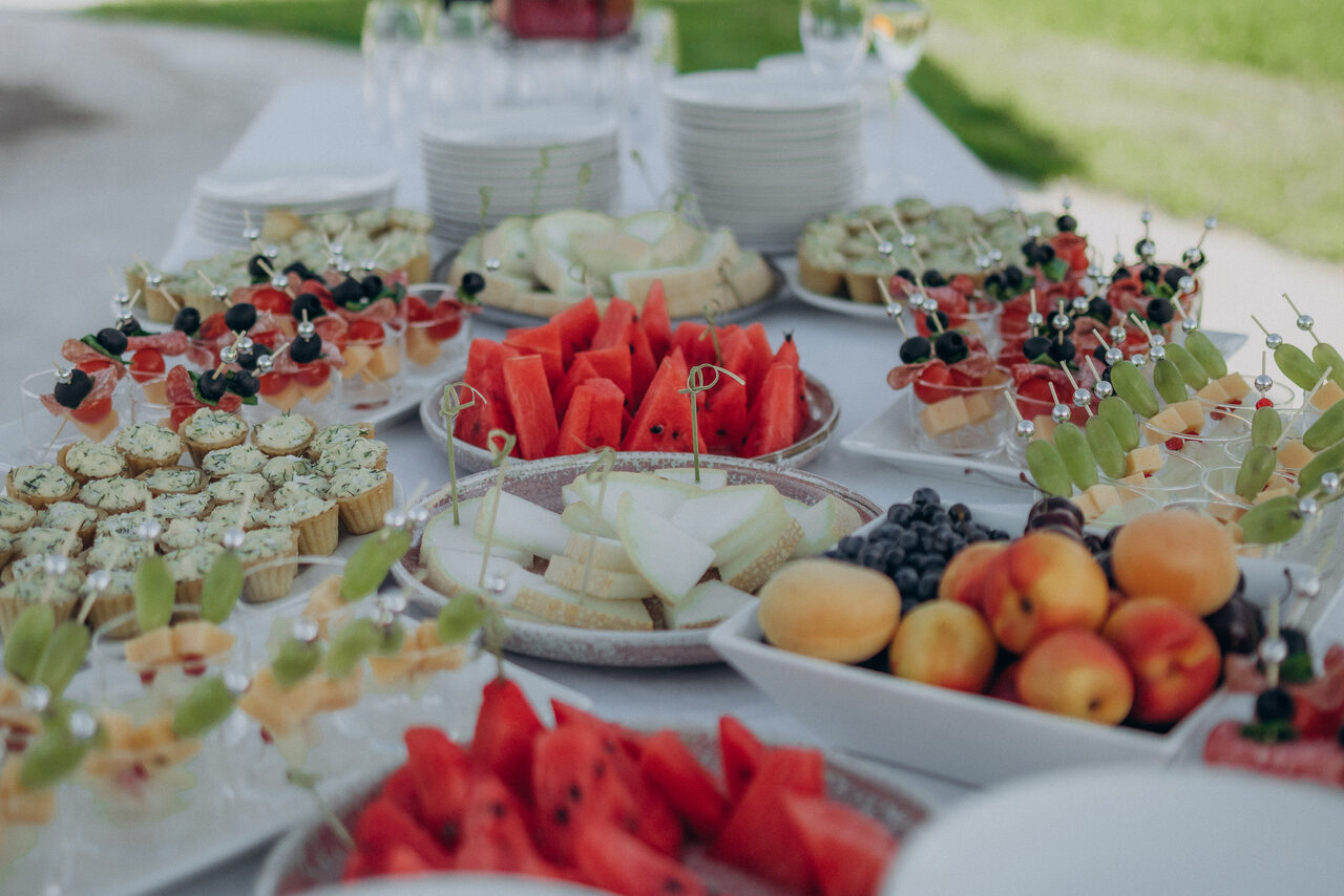 Арендовать Эко-отель "Джао Да!Ча". Шатер-Сфера. Белый стол с закусками: нарезанные фрукты, ягоды на шпажках, капкейки, десерты в стаканчиках, абрикосы, арбузы и дыни. Сложенная стопка тарелок на заднем плане. Пространство на открытом воздухе с зеленой травой.