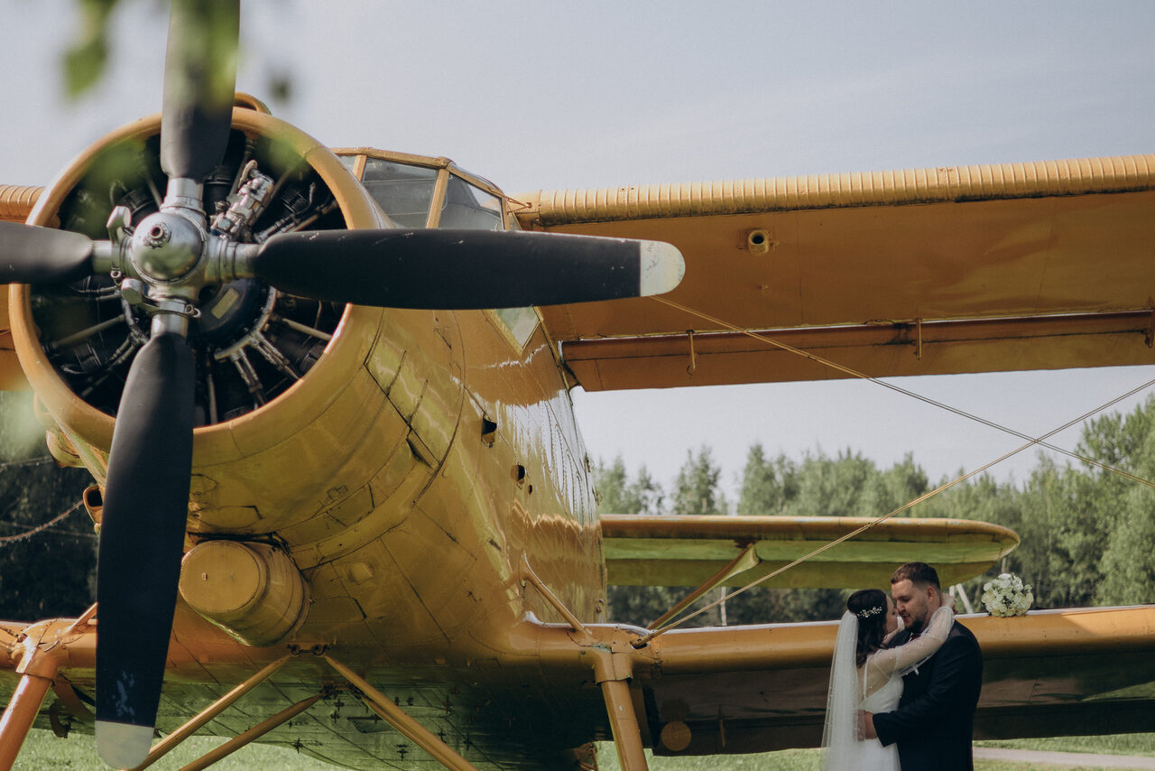 Арендовать Эко-отель "Джао Да!Ча". Шатер-Сфера. Желтый биплан на открытом воздухе с людьми возле него. Пропеллер и крыло видны крупным планом. На заднем плане деревья и голубое небо. Зеленая трава под самолетом и вокруг него.