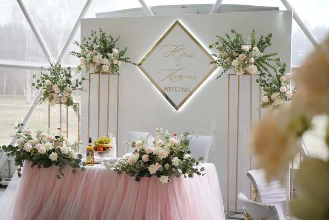 Арендовать Эко-отель "Джао Да!Ча". Шатер-Сфера. Свадебное оформление с стулом и столом, покрытым розовой тканью. На столе и вокруг высокие композиции из белых и розовых цветов и зелени. Задний фон — белая стенка с текстом "Wedding" в золотой рамке. Естественное освещение.