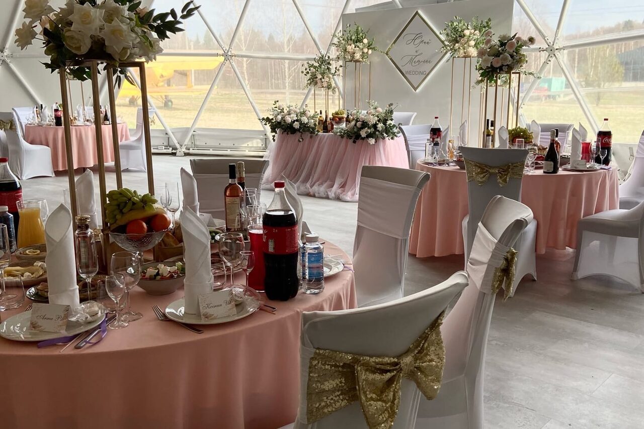 Арендовать Эко-отель "Джао Да!Ча". Шатер-Сфера. Купольное помещение с обеденными столами, покрытыми розовыми скатертями. Стулья с белыми чехлами и золотыми бантами. На столах декор из цветов, фрукты и напитки. Естественное освещение через большие окна. Панно с цветочным оформлением и надписью.