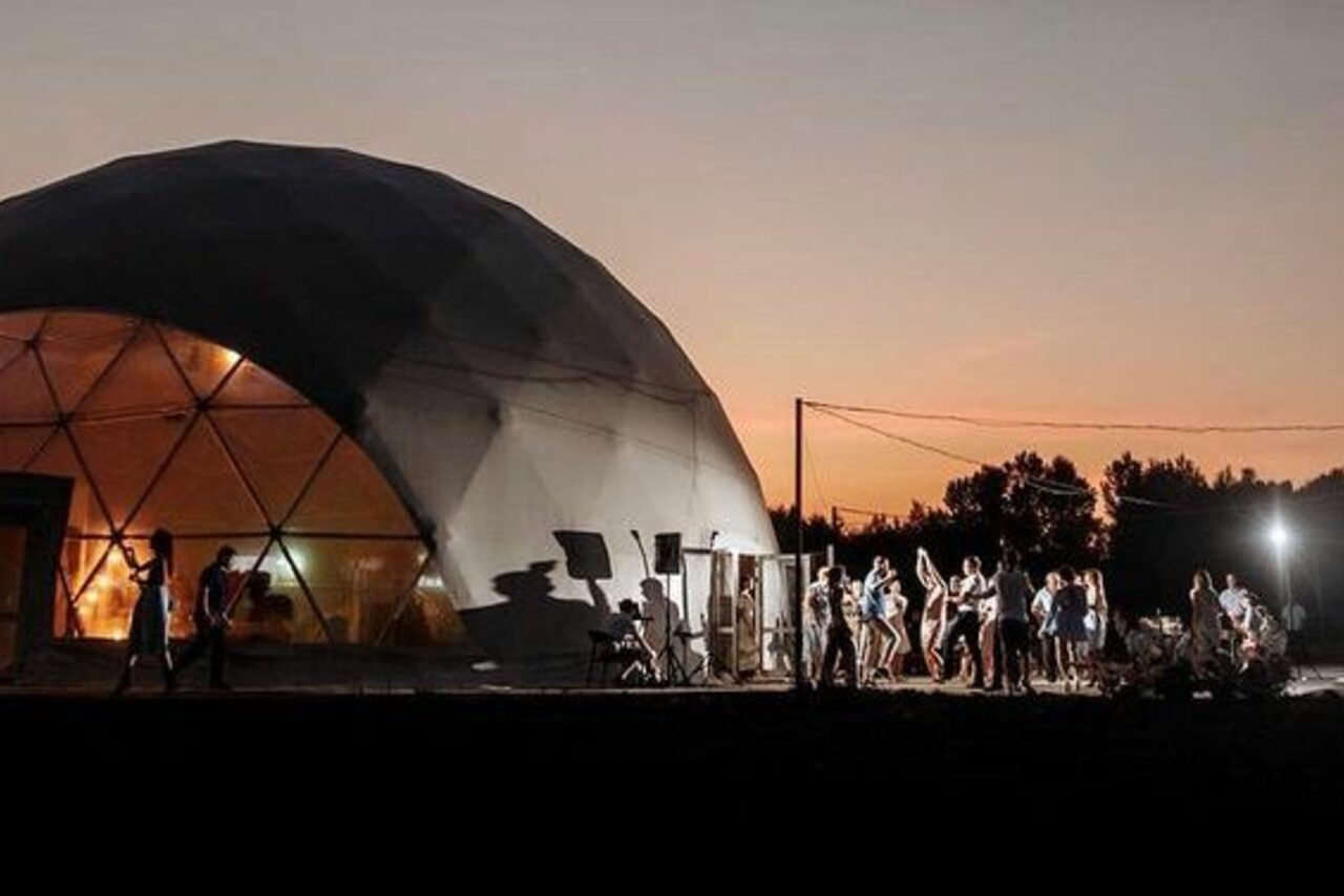 Арендовать Эко-отель "Джао Да!Ча". Шатер-Сфера. Купольная конструкция с геодезической формой, прозрачные панели на части купола, позволяющие видеть внутреннее освещение. Группа людей на улице, деревья на заднем плане, вечернее небо с закатом.