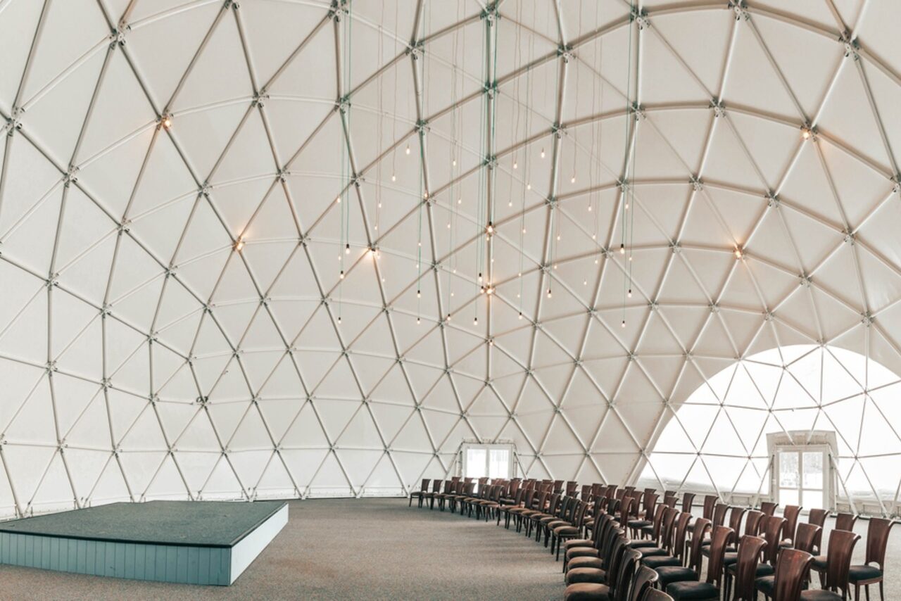 Арендовать Эко-отель "Джао Да!Ча". Шатер-Сфера. Геодезический купол с каркасом из треугольников, установлен ряд стульев для зрителей, есть небольшая сцена. Освещение представлено подвесными лампами. Цвета основные: белый купол, темно-серая сцена, коричневые стулья, серый пол.