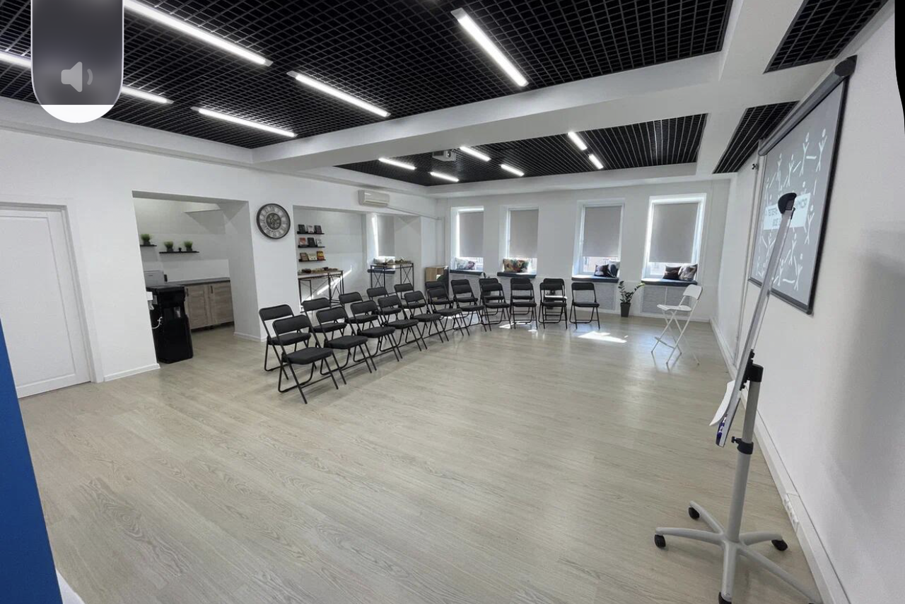 Арендовать Уютный тренинговый зал на Таганской. Современное светлое помещение с деревянным полом, расставлены черные складные стулья в два ряда. Справа — флипчарт, слева — проем в небольшую зону с раковиной и полками. Потолок с черными панелями и встроенным освещением. Белые стены, окна с жалюзи.