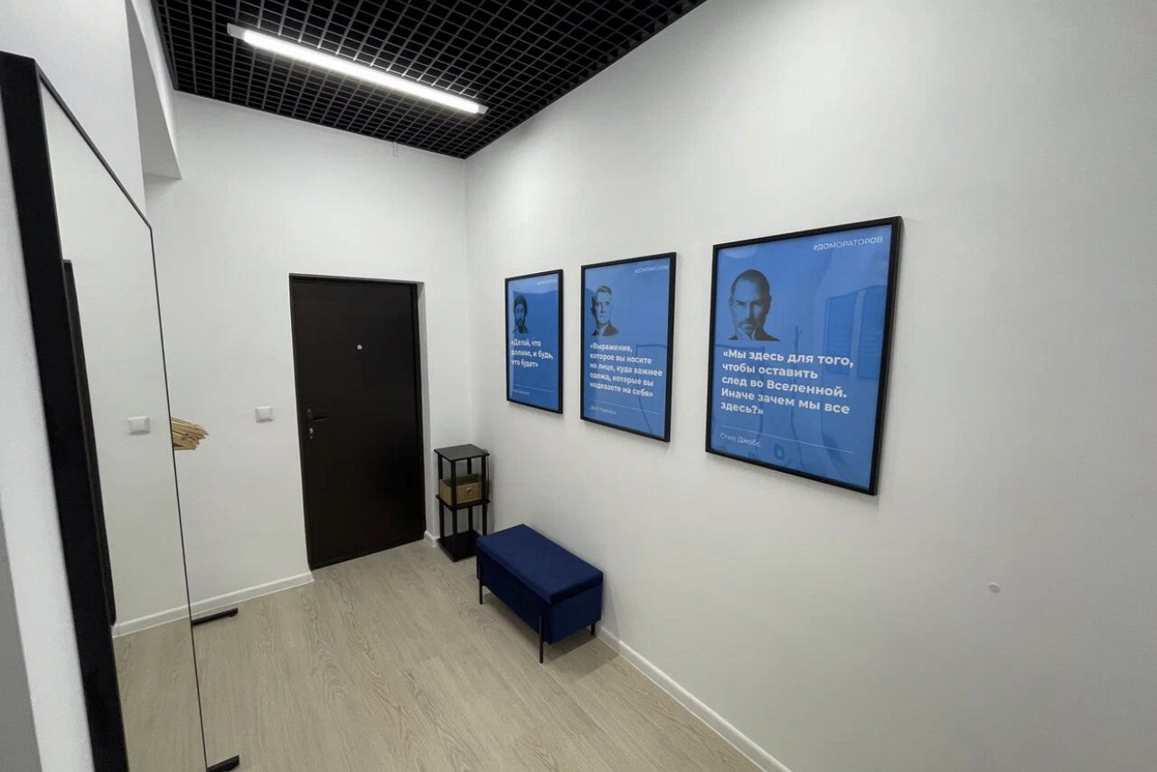 Арендовать Уютный тренинговый зал на Таганской. Узкий коридор с белыми стенами и светлым деревянным полом. Черный подвесной потолок с встроенным светильником. Три синие картины на стене, черная дверь, синяя скамья и черный декор на полу.