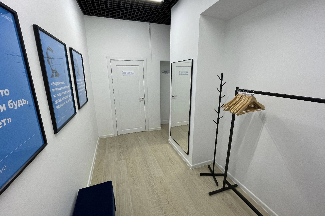 Арендовать Уютный тренинговый зал на Таганской. Узкое пространство с белыми стенами, светлым деревянным полом и черным подвесным потолком. Справа зеркало, черная стойка с вешалками и лавка. Слева три синих плаката в рамках. Две белые двери с надписями.