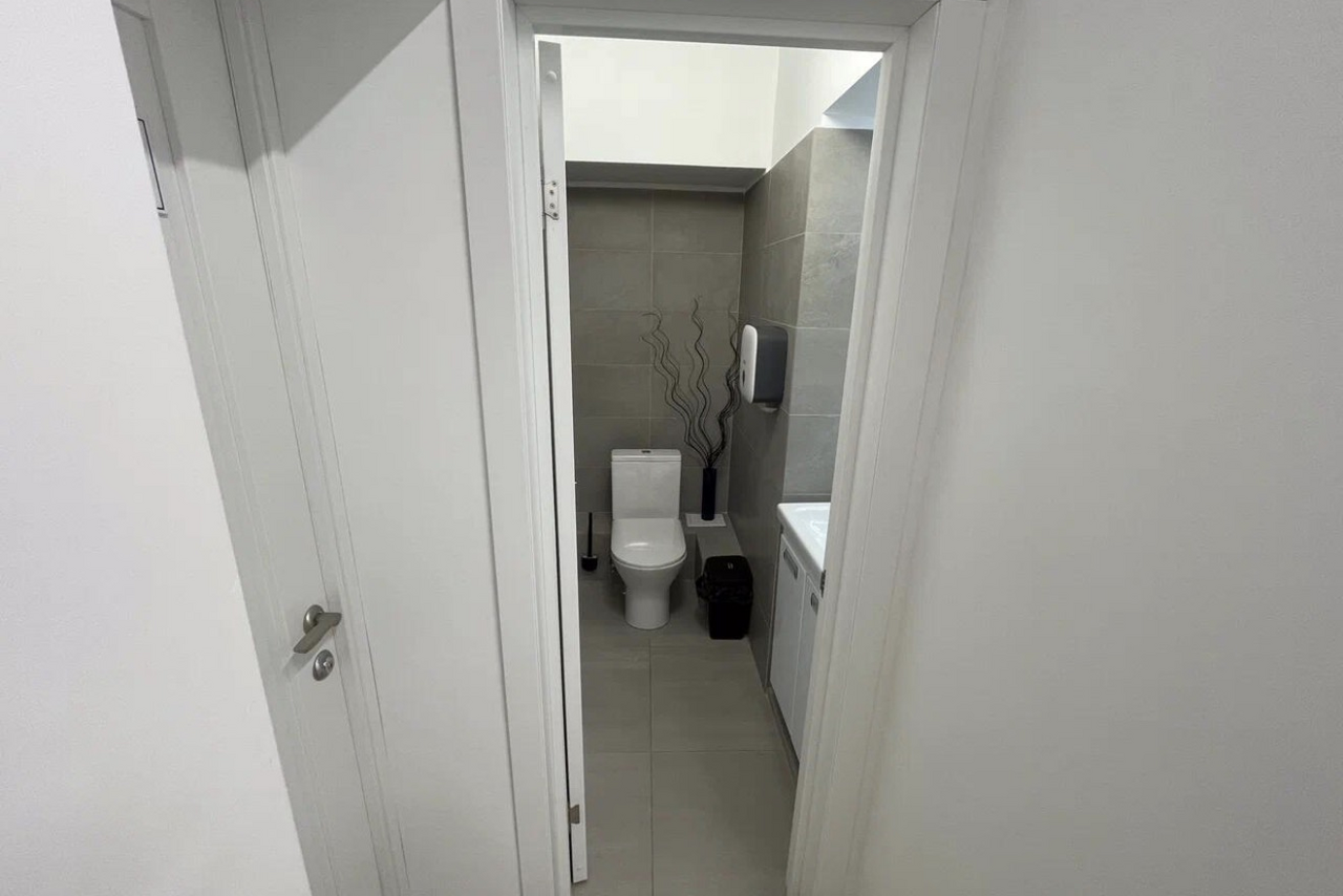 Арендовать Уютный тренинговый зал на Таганской. Дверь ведет в санитарную комнату с унитазом, белой раковиной и диспенсером для мыла. Бежевые стены и пол из плитки, белая дверь и потолок. Черная корзина для мусора и декоративные элементы рядом с унитазом.