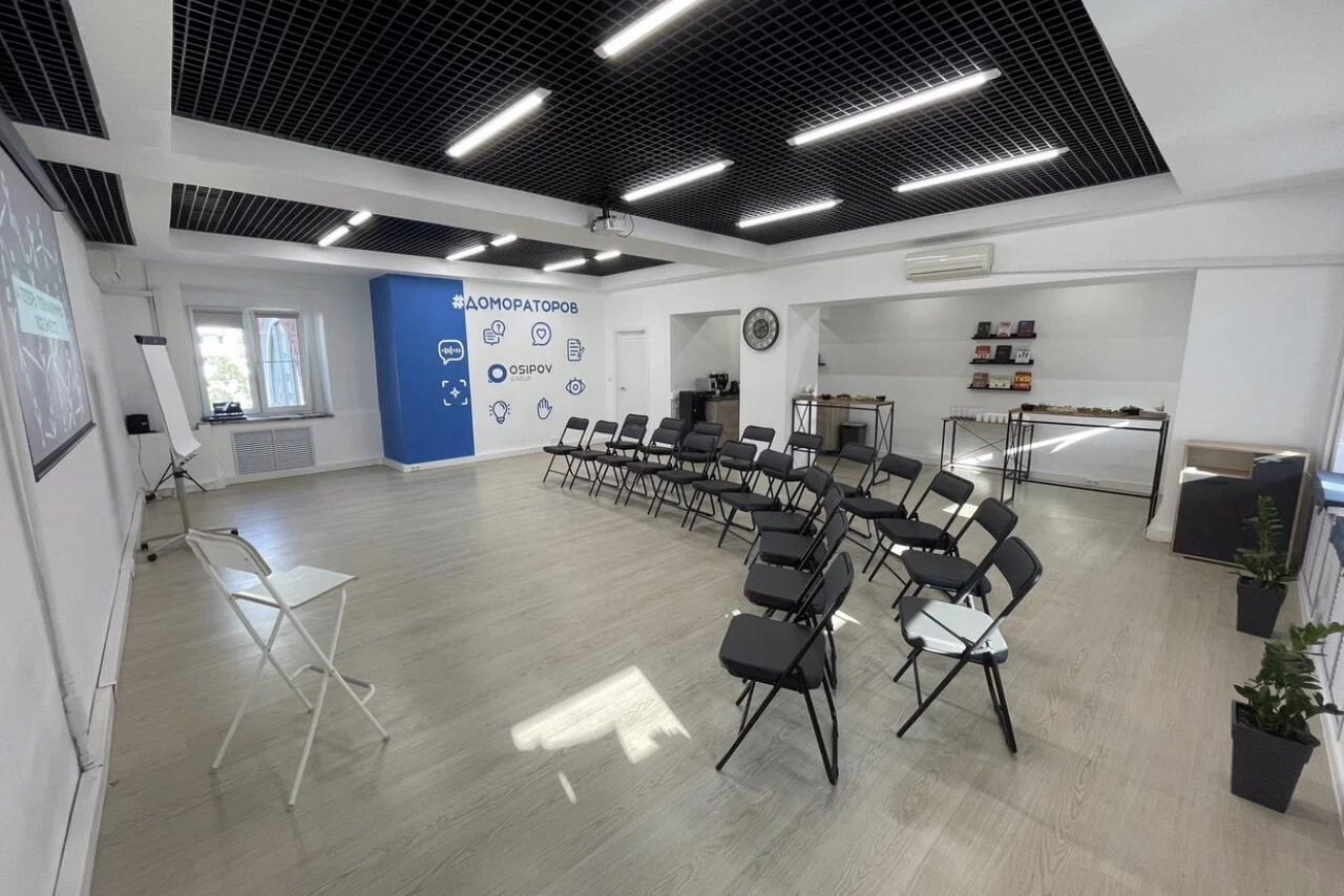 Арендовать Уютный тренинговый зал на Таганской. Просторное помещение с двумя рядами черных стульев, белыми стенами и деревянным полом. На стене виден экран и флипчарт. В правом углу столы с предметами, слева у окна кондиционер. Потолок черно-белый с подвесными светильниками.