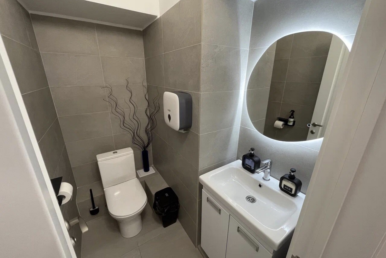 Арендовать Уютный тренинговый зал на Таганской. Туалетная комната с серыми плиточными стенами и полом. Унитаз, держатель для туалетной бумаги, щетка. Умывальник с белой тумбой и круглым зеркалом с подсветкой. Диспенсер для мыла, бумажный диспенсер, черное мусорное ведро, ваза с декоративными ветками.