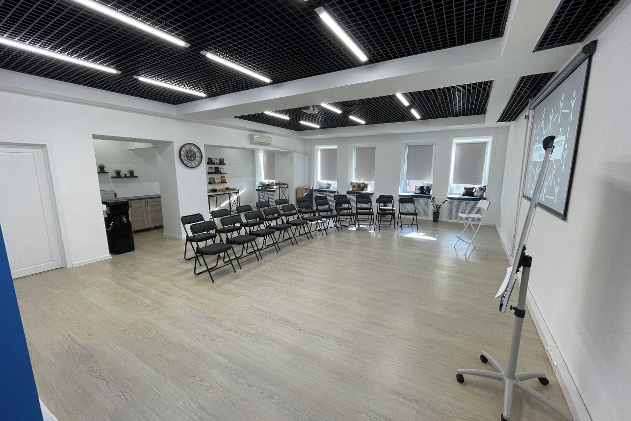 Арендовать Уютный тренинговый зал на Таганской. Просторное помещение с белыми стенами и светлым деревянным полом, черный ячеистый потолок. Ряды черных складных стульев. Справа – флипчарт на подставке. В глубине встроенная кухня, полки, белый складной стол, окна с белыми шторами.