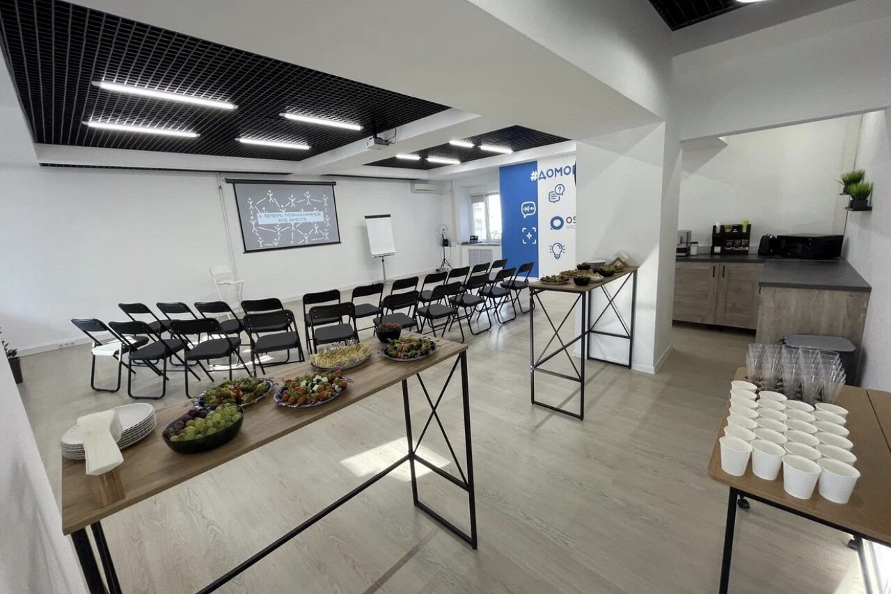 Арендовать Уютный тренинговый зал на Таганской. Просторная комната с черными складными стульями, расположенными в ряд. На стене проекционный экран и флипчарт. Два стола с закусками и напитками. Светлое деревянное покрытие пола. Потолок с черной решетчатой структурой и встроенными светильниками. Стена с синим баннером.