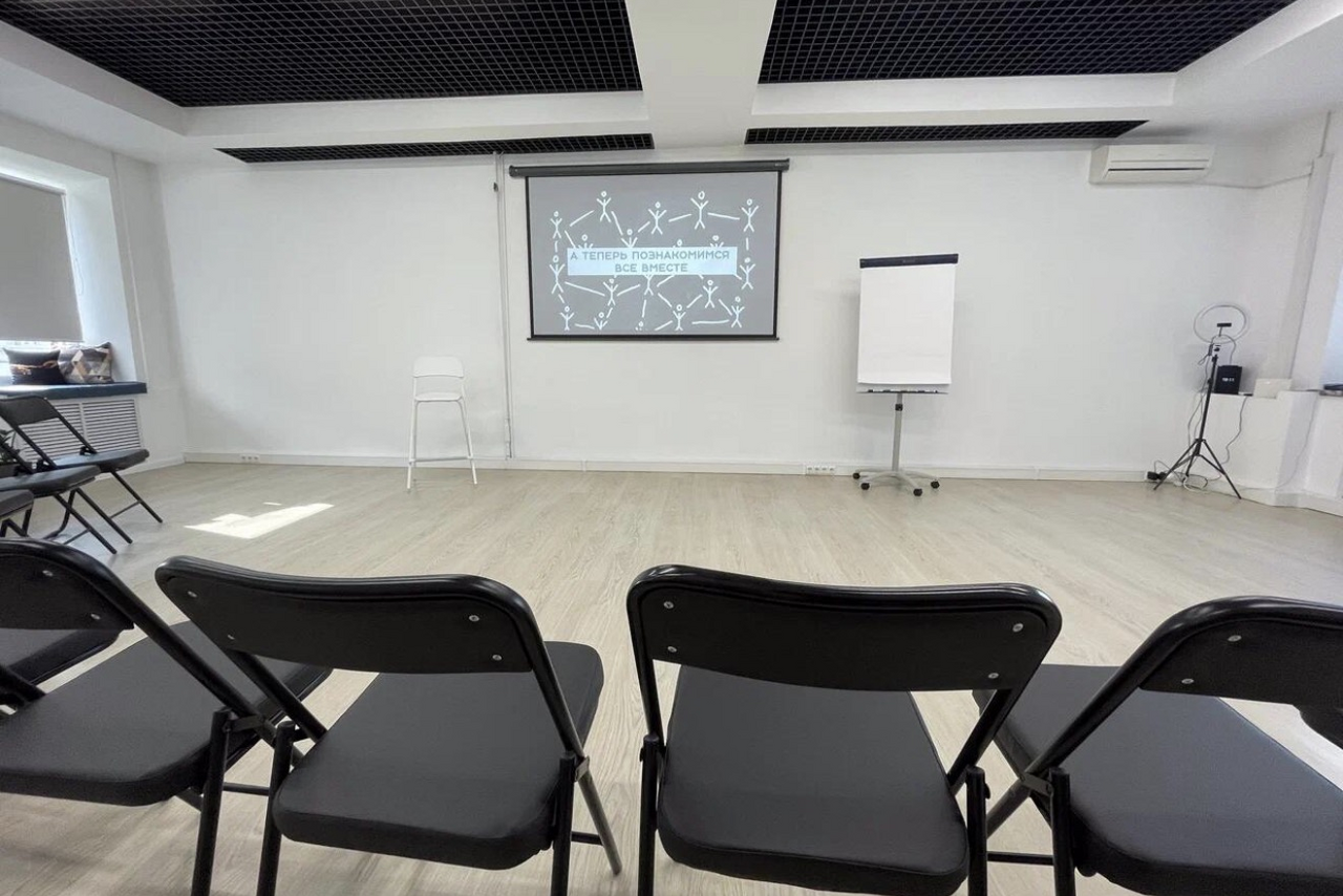 Арендовать Уютный тренинговый зал на Таганской. Просторное помещение с деревянным светлым полом, белыми стенами и черным потолочным грилем. Ряды черных стульев, экран с проектором, флипчарт на подставке, белый стул у стены. Свет из большого окна справа, кондиционер на стене.