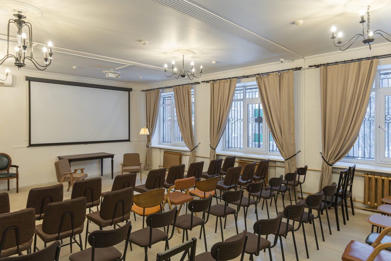 Арендовать Open Space (Открытое пространство) для мероприятий. Помещение с рядами стульев, проекторным экраном, столом и креслами. Пастельные серые и коричневые оттенки, паркетный пол. Окна с бежевыми шторами и решетками, люстры на потолке, радиаторы под окнами.