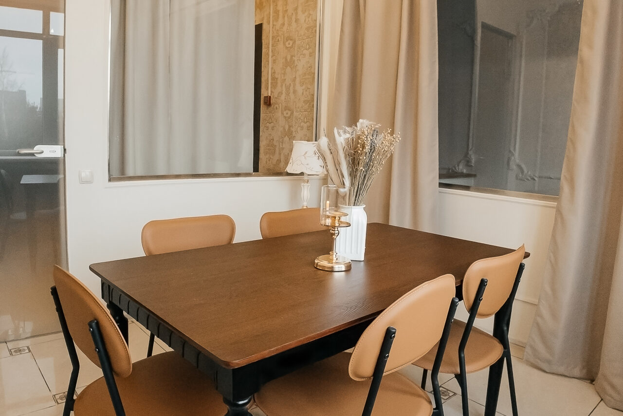 Арендовать Open Space (Открытое пространство) для мероприятий. Прямоугольный деревянный стол с шестью коричневыми стульями на металлических ножках. На столе декоративная композиция в вазе. Светлый интерьер с бежевыми шторами и светлыми стенами. Пол из плитки. Видно дверь и окно.