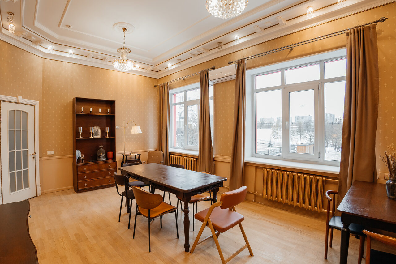 Арендовать Open Space (Открытое пространство) для мероприятий. Прямоугольная комната с высокими потолками, светлыми стенами и деревянным полом. В центре деревянный стол и стулья. Справа окно с занавесками, под ним батарея. Слева шкаф и настольная лампа. Встроенные потолочные светильники и подвесные люстры.