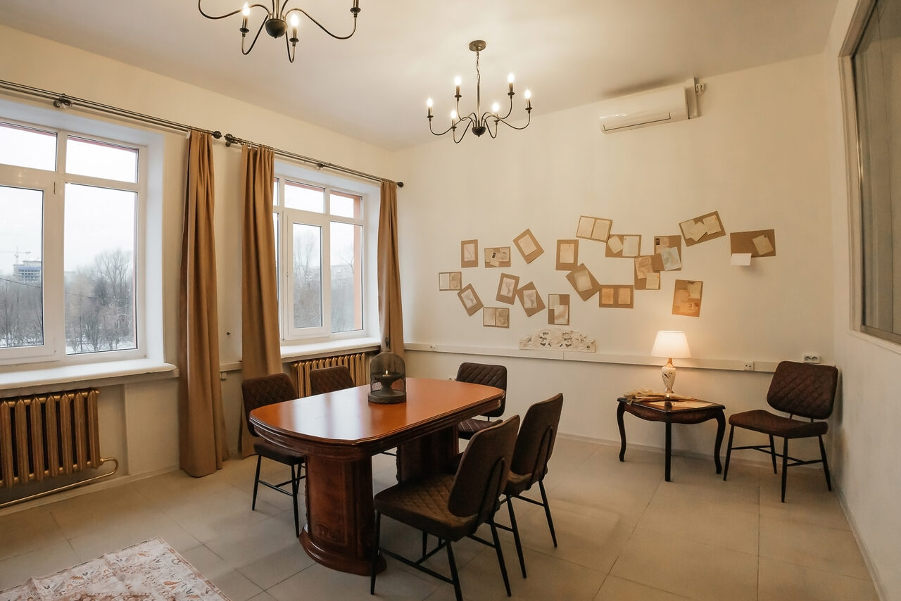 Арендовать Open Space (Открытое пространство) для мероприятий. Просторная комната с двумя окнами, столом с четырьмя стульями, двумя креслами и тумбочкой с лампой. На стене декор из пустых рамок. Светлые шторы, люстра и настенные светильники. Белые стены, бежевый пол. Радиатор под окнами. Кондиционер над картинками.