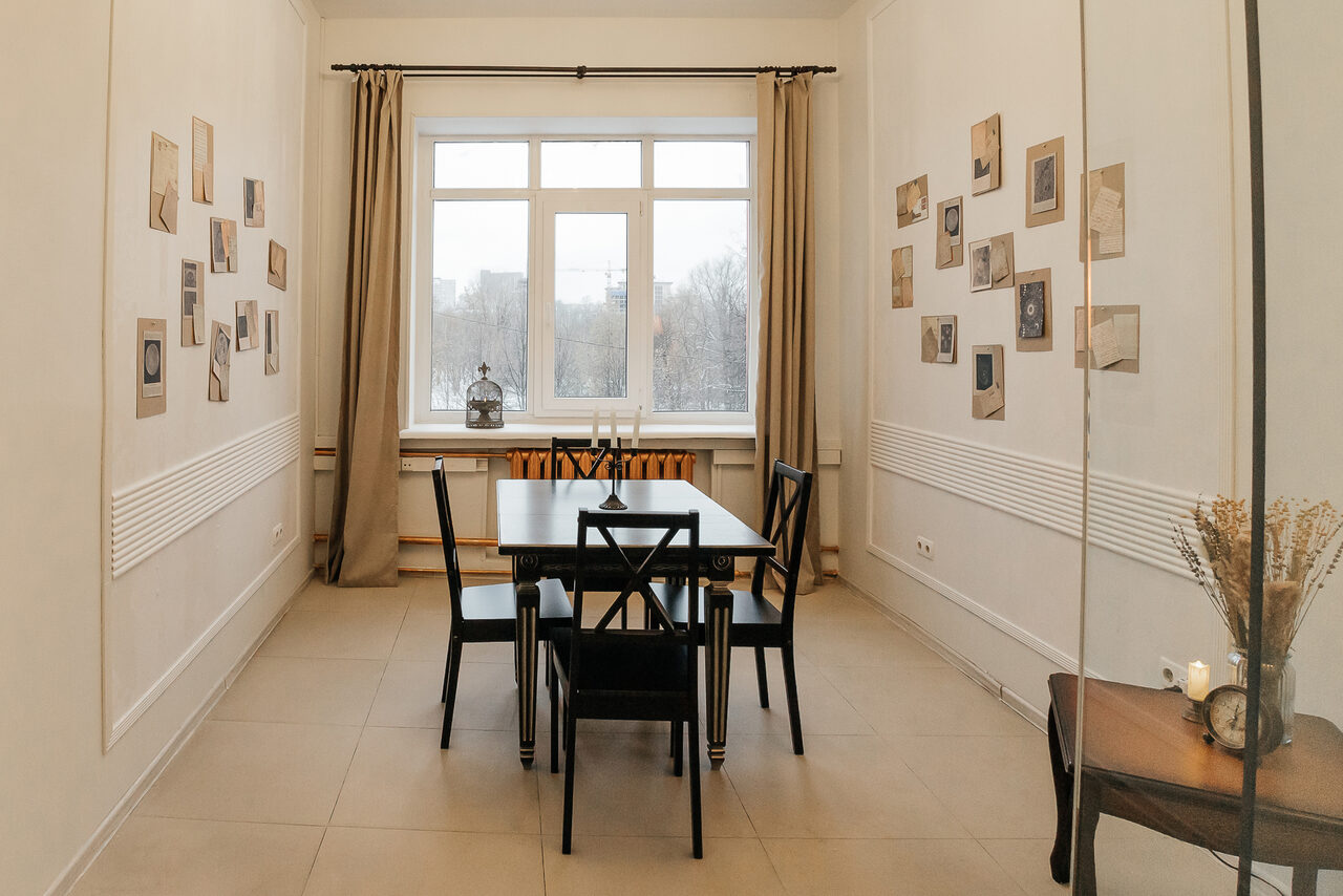 Арендовать Open Space (Открытое пространство) для мероприятий. Прямоугольная комната с высокими потолками. По центру деревянный стол и четыре стула. Светлая плитка на полу, белые стены украшены фотографиями. На переднем плане столик с вазой. Большое окно с бежевыми шторами.