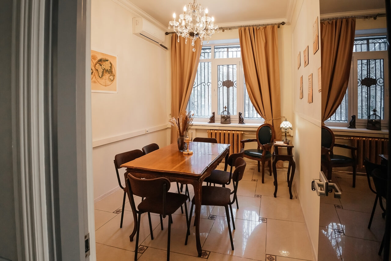 Арендовать Open Space (Открытое пространство) для мероприятий. Небольшая комната с деревянным столом и шестью стульями, люстра, светлые стены, картины, окна с металлическими решетками, шторы, настольная лампа, плиточный пол. Пространство аккуратное и светлое.